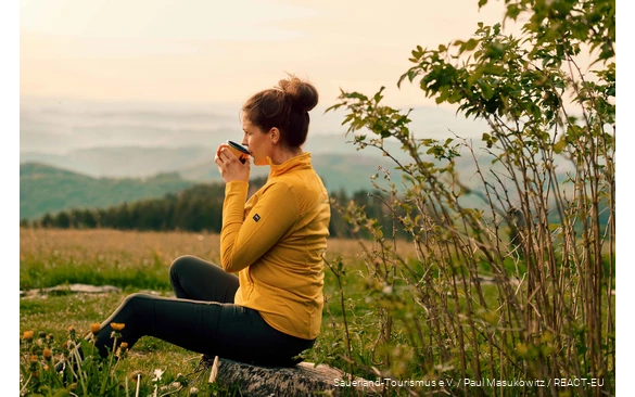 Eine Wanderin genießt ihre Rast bei einer Tasse Kaffee und Blick über die Landschaft.