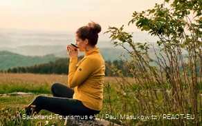 Eine Wanderin genießt ihre Rast bei einer Tasse Kaffee und Blick über die Landschaft.